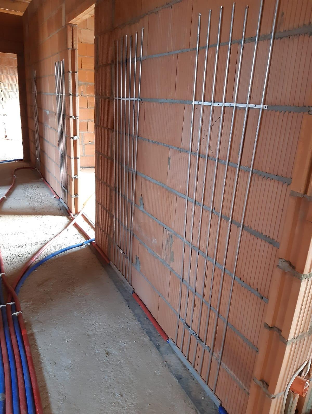Instalacja rur grzewczych w ścianie z pustaków ceramicznych, widoczne metalowe wsporniki i fragmenty rur w podłodze.
