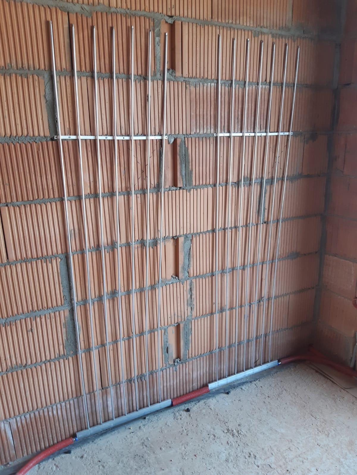 Montaż pionowych rurek ogrzewania podłogowego ściennego na ceglanej ścianie, widoczne poziome wsporniki i fragment instalacji w podłodze.
