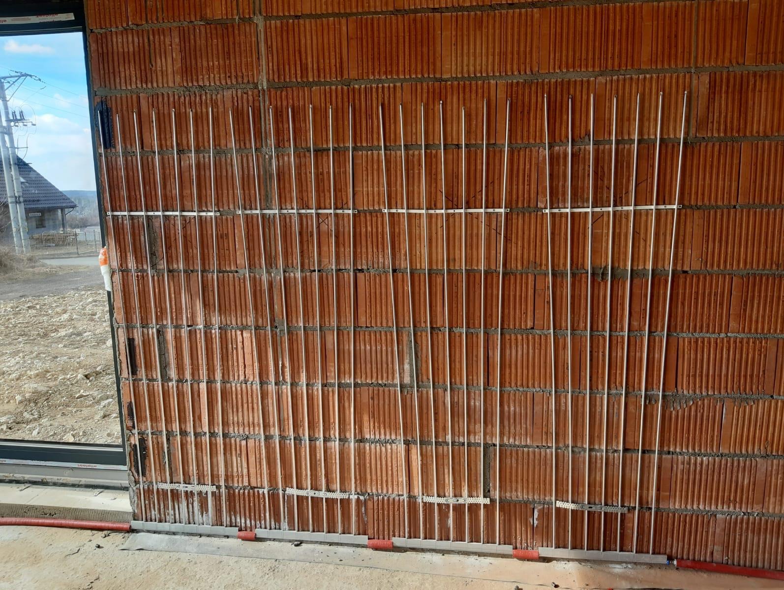 Montaż pionowych rurek ogrzewania podłogowego na ceglanej ścianie, widoczne okno i fragment krajobrazu za nim.