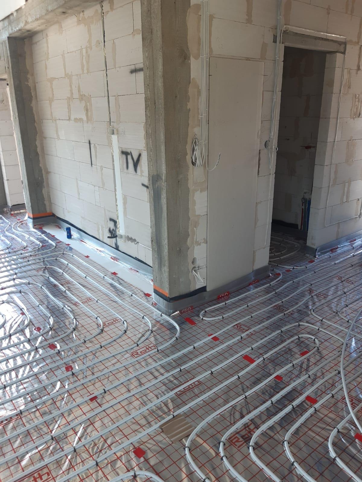 Białe rury ogrzewania podłogowego ułożone na folii z czerwoną siatką w surowym wnętrzu budynku z bloczków betonowych.