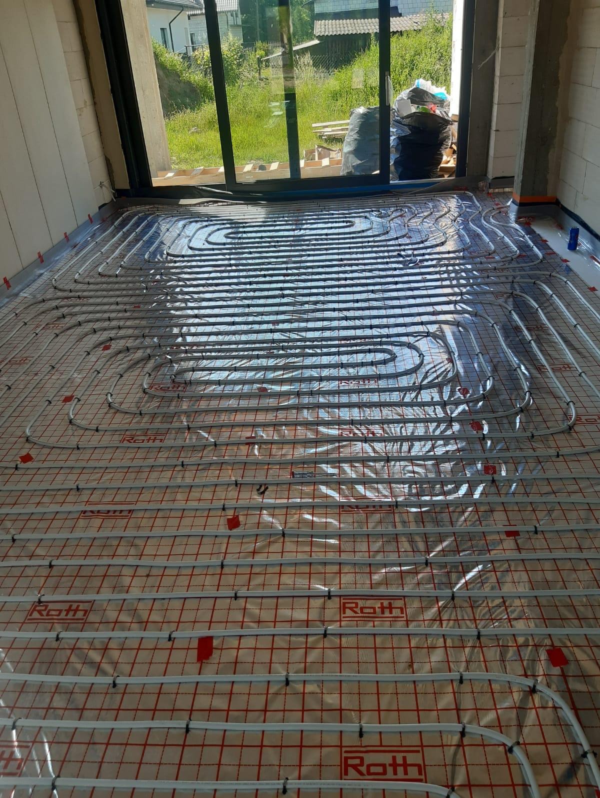 Rozłożona instalacja ogrzewania podłogowego Roth na folii z rastrem w nowym budynku, widoczne rury grzewcze i mocowania, w tle okno z widokiem na zieleń.