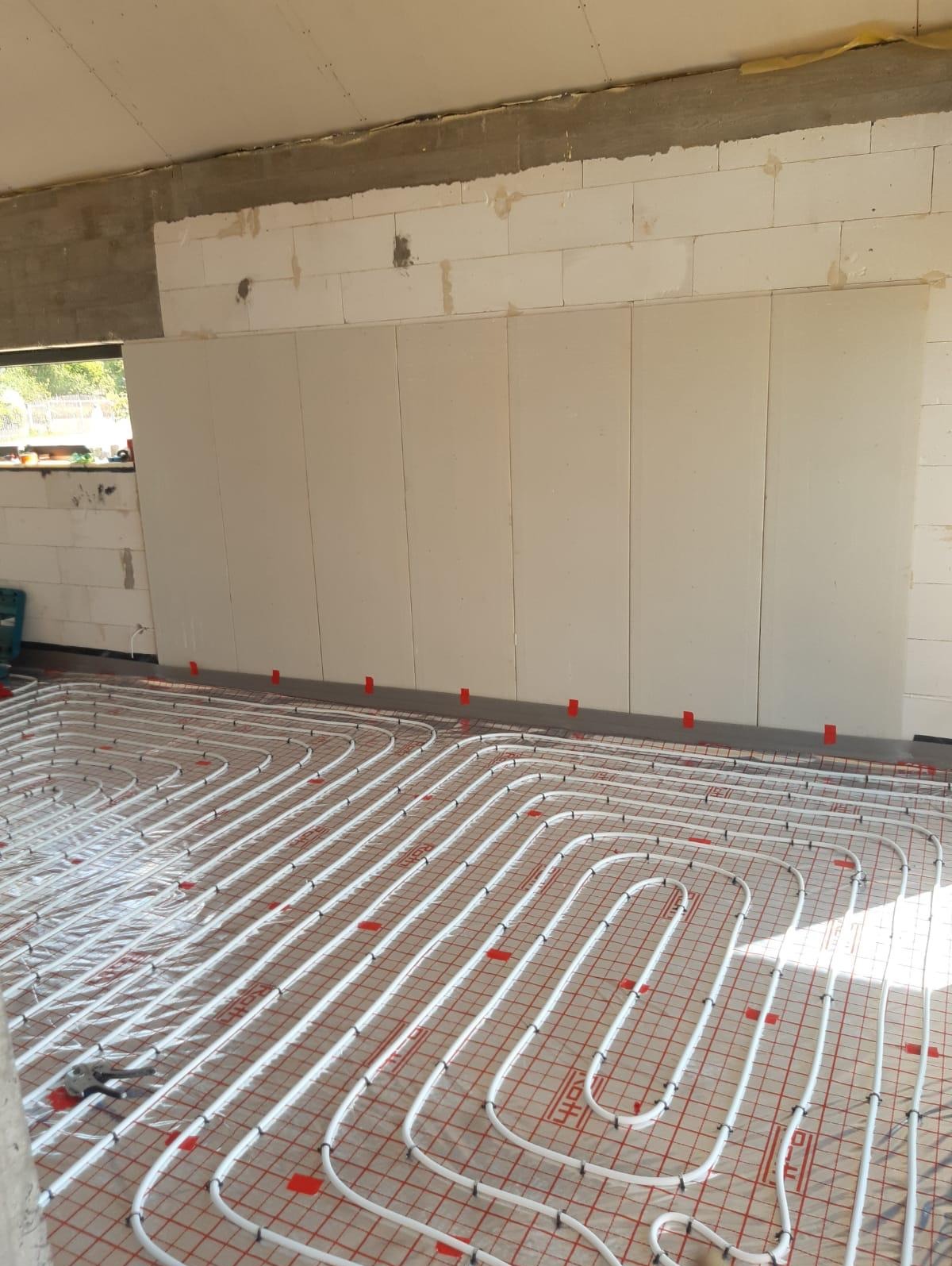 Białe rury systemu ogrzewania podłogowego ułożone na czerwonej siatce w surowym wnętrzu budynku w trakcie instalacji.