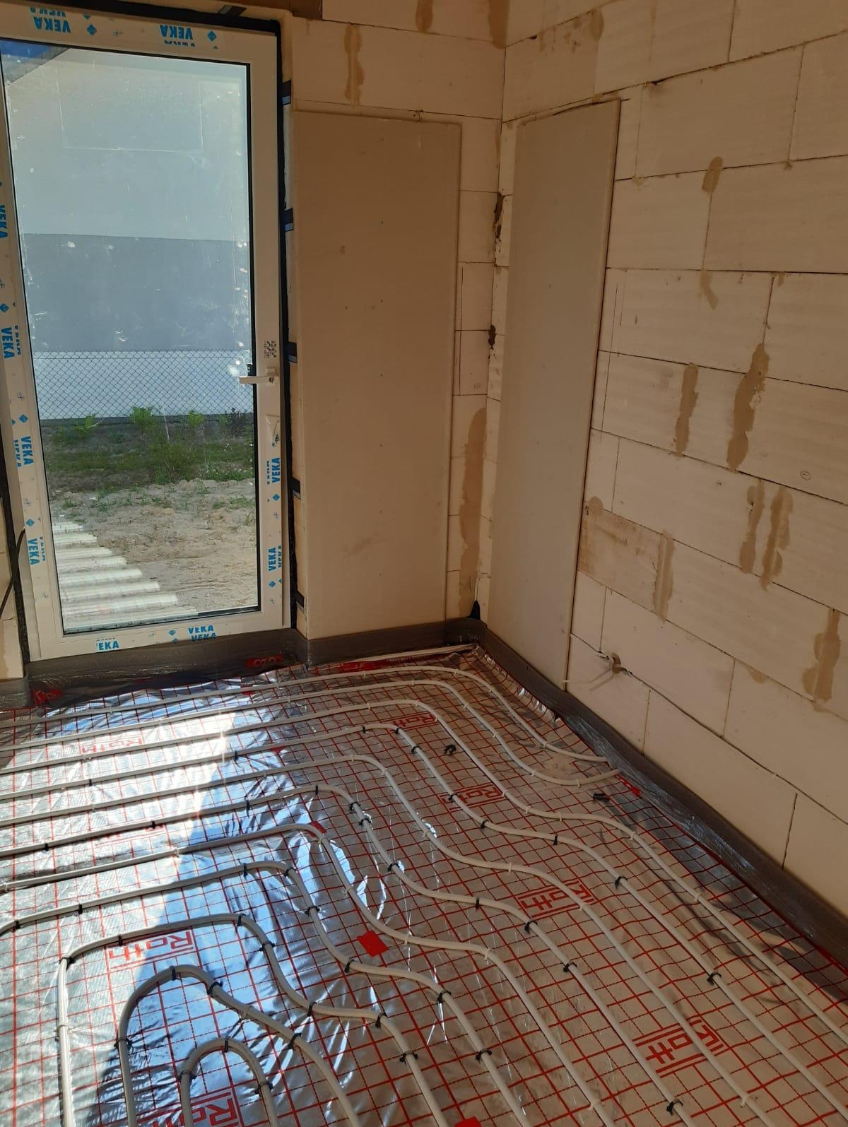 Białe rury ogrzewania podłogowego ułożone na czerwonej macie w surowym wnętrzu, widok przez okno na zewnątrz.