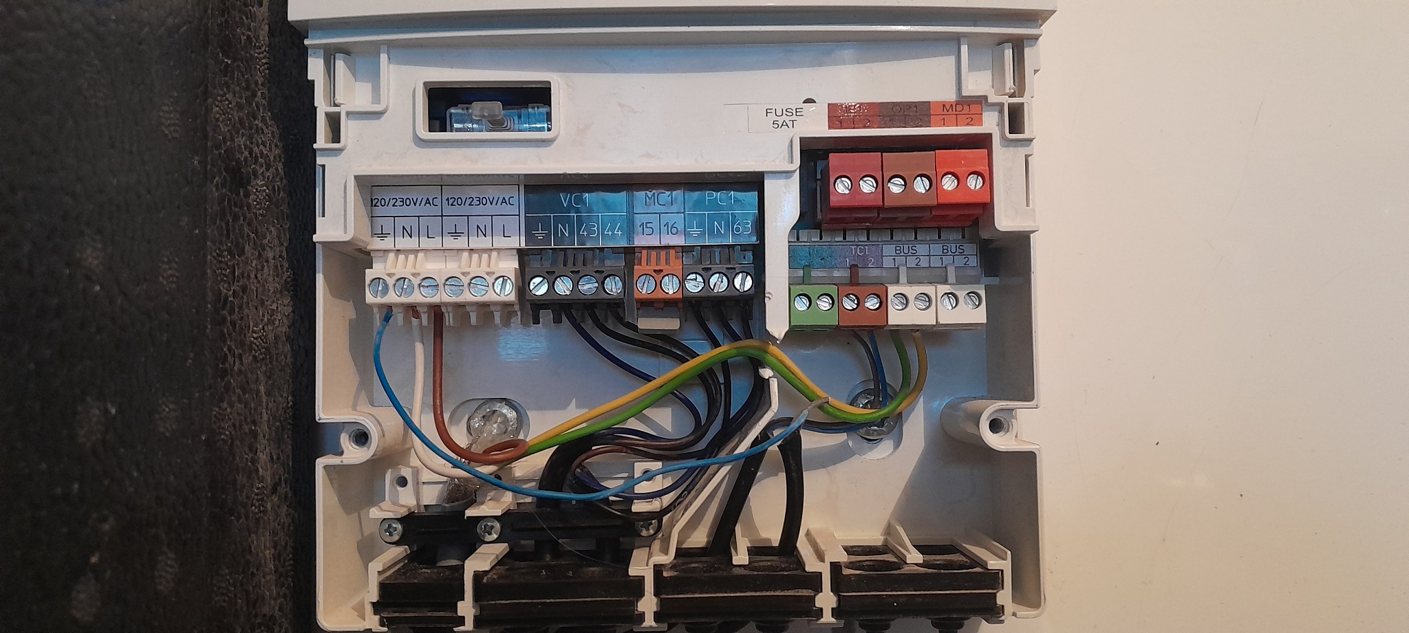 Otwarta skrzynka elektryczna z podłączonymi przewodami o różnych kolorach (niebieski, brązowy, czarny, zielono-żółty) do listew zaciskowych z oznaczeniami napięcia i numerami, widoczny bezpiecznik...