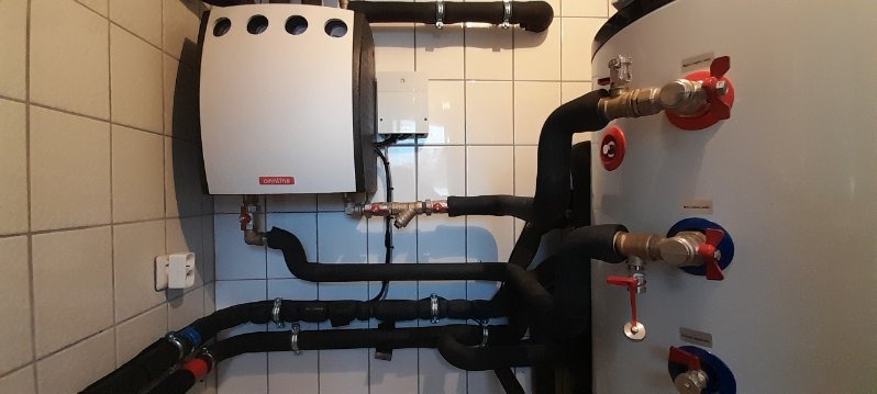 Biały piec grzewczy z podłączeniami hydraulicznymi i zbiornikiem na wodę w kotłowni wyłożonej białymi płytkami.