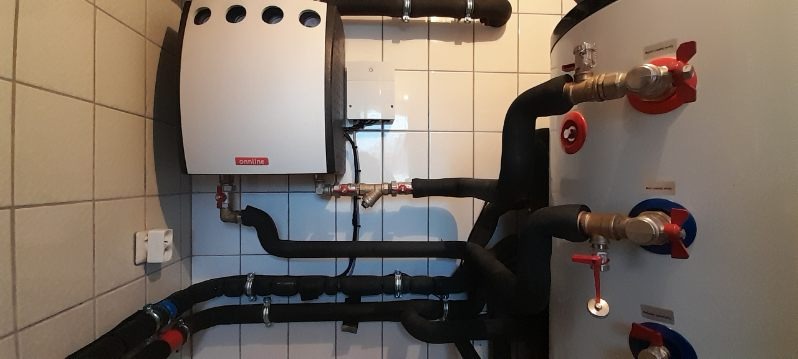 Biały piec gazowy zamontowany na ścianie z ceramicznych płytek w kotłowni, z widocznymi rurami, zaworami i zbiornikiem akumulacyjnym.