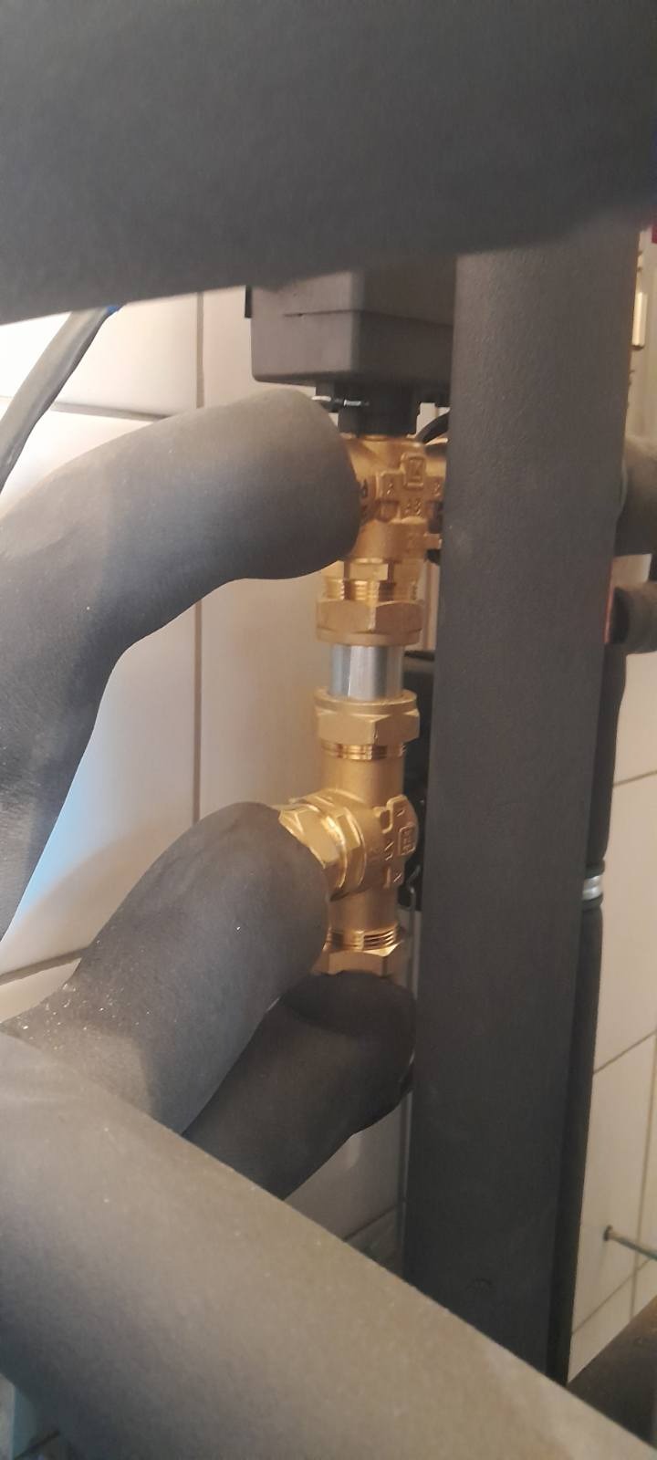 Złącze hydrauliczne z brązowymi zaworami i izolowanymi rurami, widoczne na tle białych płytek.