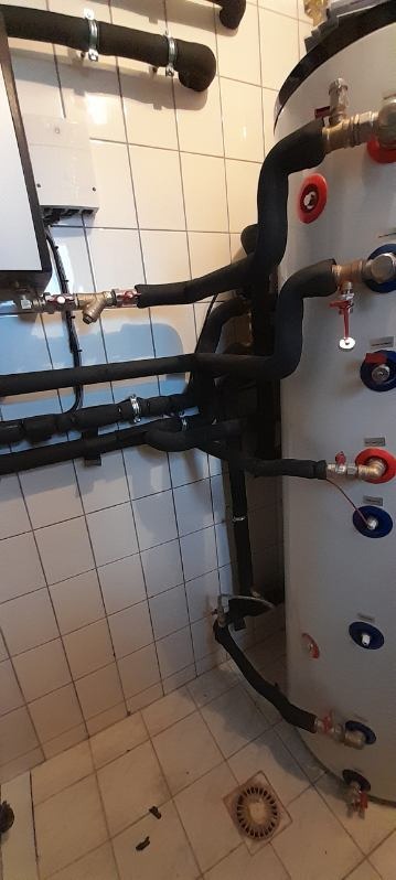 Bojler z podłączonymi rurami w izolacji, widoczne zawory z czerwonymi i niebieskimi pokrętłami, instalacja w pomieszczeniu z wyłożonymi kafelkami ścianami i podłogą.