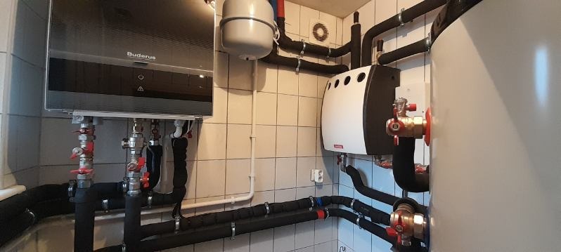 Kotłownia z piecem gazowym Buderus, zasobnikiem ciepłej wody i rozbudowanym systemem rur w pomieszczeniu wyłożonym kafelkami.
