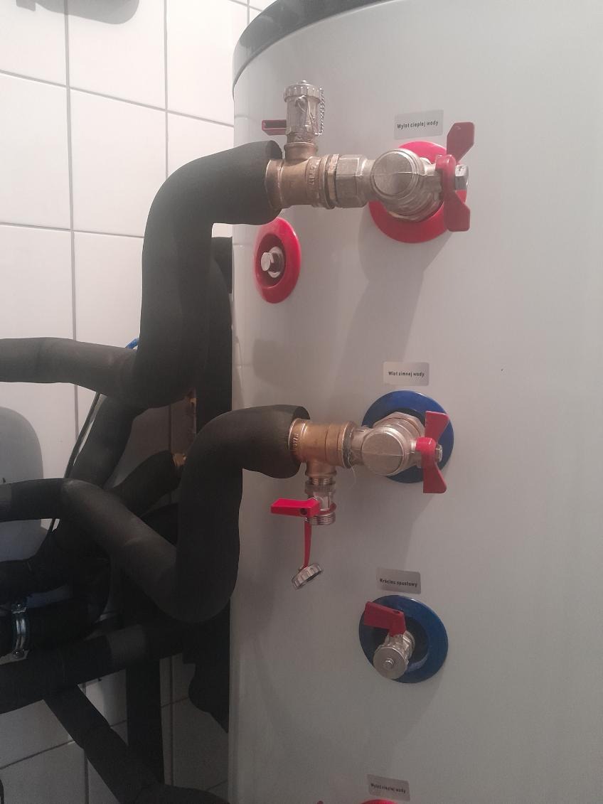 Instalacja zaworów i rur w kotłowni z widocznym zasobnikiem ciepłej wody, izolowane rury, zawory z czerwonymi i niebieskimi pokrętłami, białe płytki na ścianie.