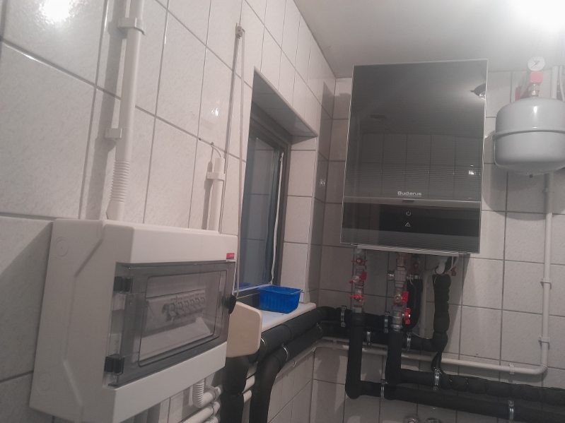 Nowoczesny piec gazowy marki Buderus zamontowany na ścianie z białych płytek, widoczna instalacja hydrauliczna z czarnymi rurami i czerwonymi zaworami oraz skrzynka elektryczna z bezpiecznikami.