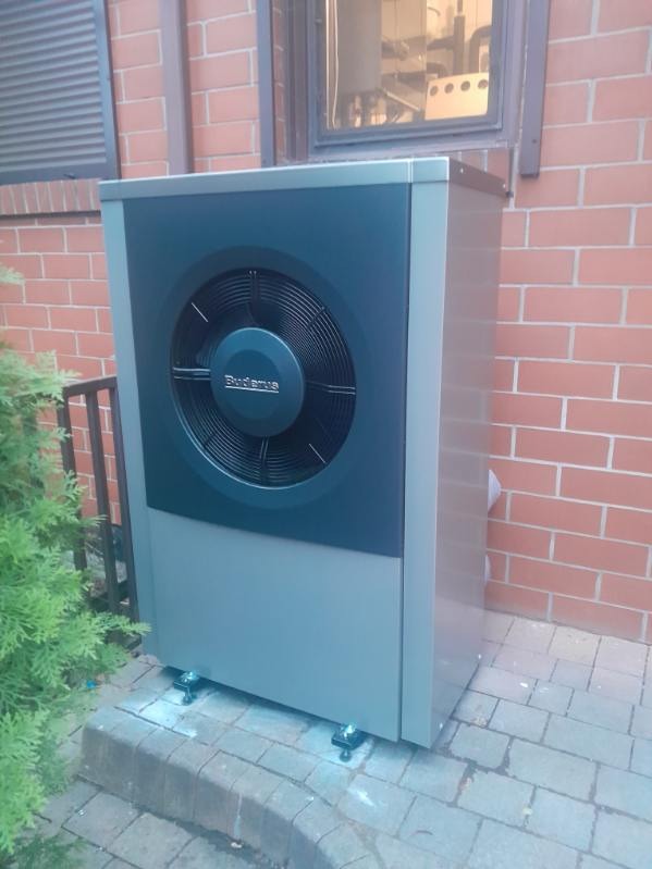 Szara pompa ciepła Buderus zainstalowana przy ceglanym budynku, widoczna z boku, z zielonym krzewem po lewej stronie i oknem w tle.