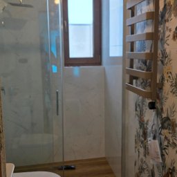 Drex - Wykończona łazienka z kabiną prysznicową, wiszącą toaletą, grzejnikiem drabinkowym na ścianie w kwiaty. Na podłodze panele imitujące drewno. Widoczny stołek roboczy.