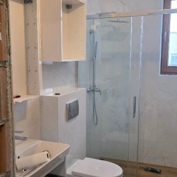 Drex - Nowoczesna łazienka w trakcie wykańczania: białe meble, kabina prysznicowa, drewnopodobne panele. Widoczne narzędzia i plany na podłodze. Oświetlenie górne.