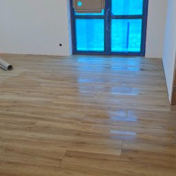 Ukończona podłoga z paneli imitujących drewno w jasnym wnętrzu. Okno zabezpieczone folią, widoczne przygotowania do dalszych prac wykończeniowych.