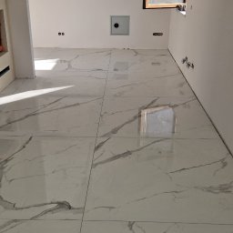 Błyszcząca, biała podłoga z marmurowym wzorem, ułożona z dużych płytek. Widoczne okno i fragment ściany z instalacją elektryczną. Minimalistyczne wnętrze w trakcie wykańczania.