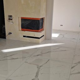 Wykończony kominek z gładzią w beżowym odcieniu, zabezpieczony folią. Na podłodze białe płytki imitujące marmur. Widoczne gniazdka elektryczne i fragment instalacji.