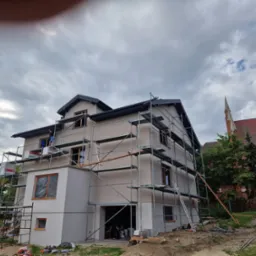 Dwupiętrowy dom w trakcie ocieplania, otoczony rusztowaniem, z widocznymi fragmentami kościoła w tle.