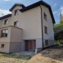 Nowo otynkowany, dwukondygnacyjny dom z garażem w piwnicy, jasna elewacja z ciemną podmurówką i stolarką okienną, widok na kościół w tle.