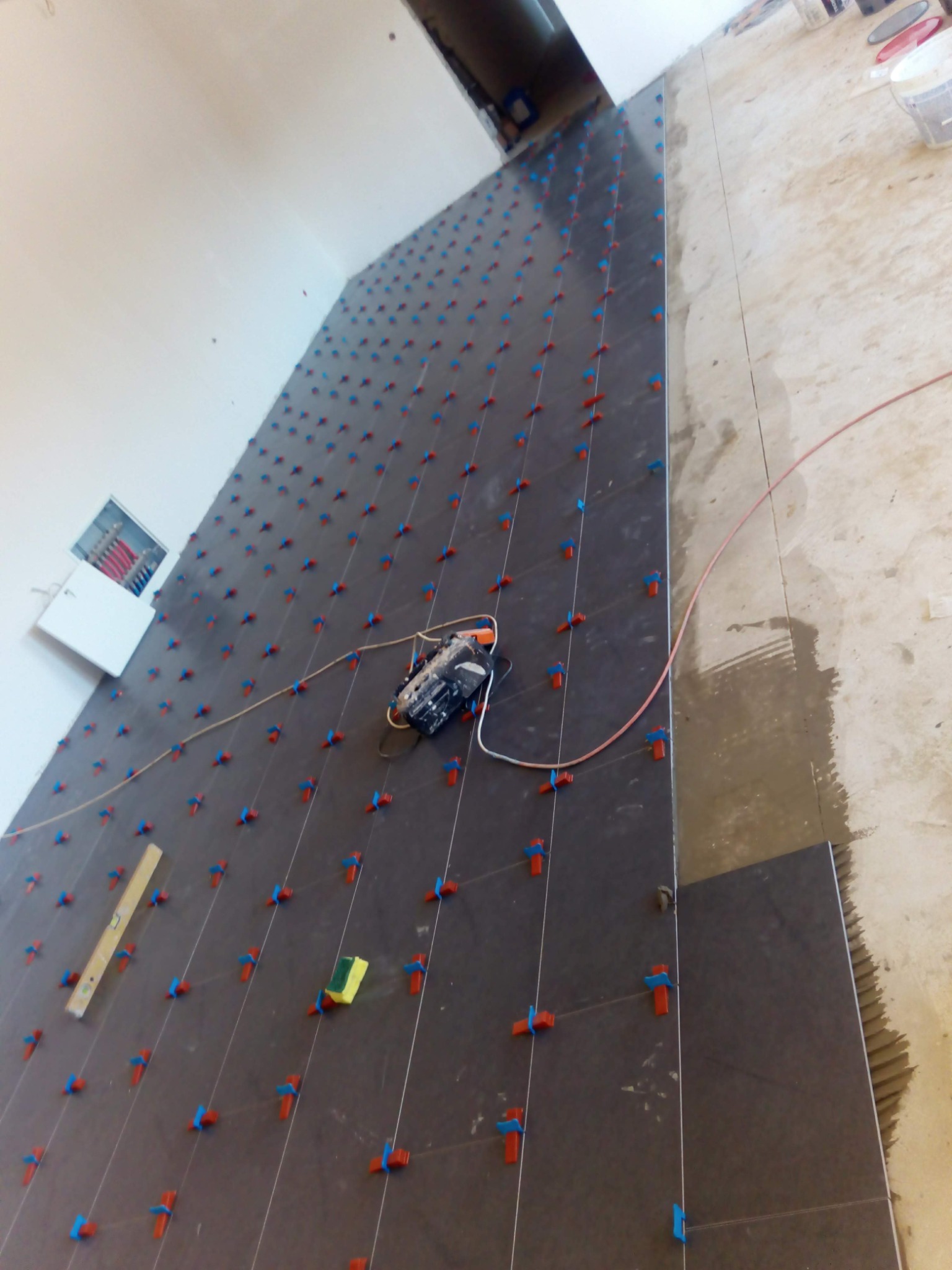 Układanie ciemnych płytek podłogowych z użyciem systemu poziomowania, widoczna poziomica, gąbka i szlifierka kątowa.