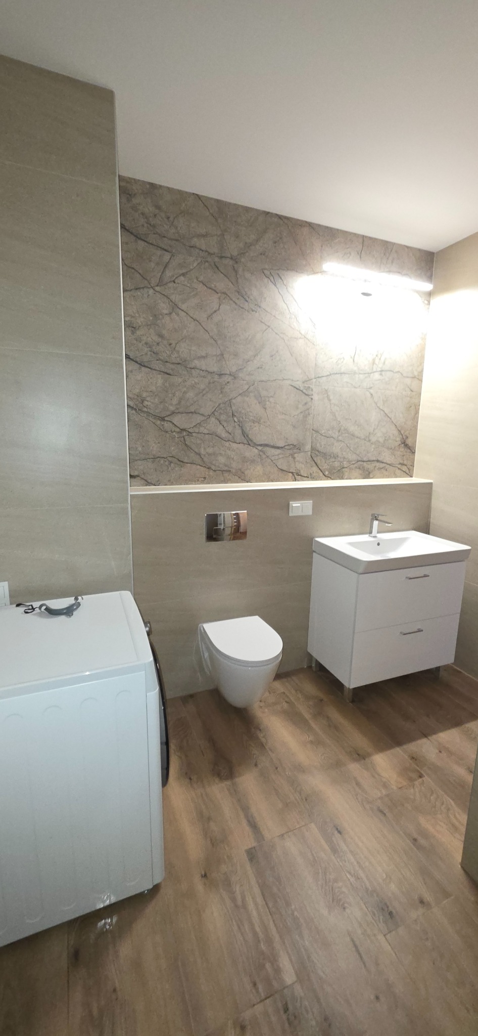 Nowoczesna łazienka z drewnopodobną podłogą, marmurową ścianą i białą armaturą. Pralka częściowo widoczna po lewej stronie. Minimalistyczny design.