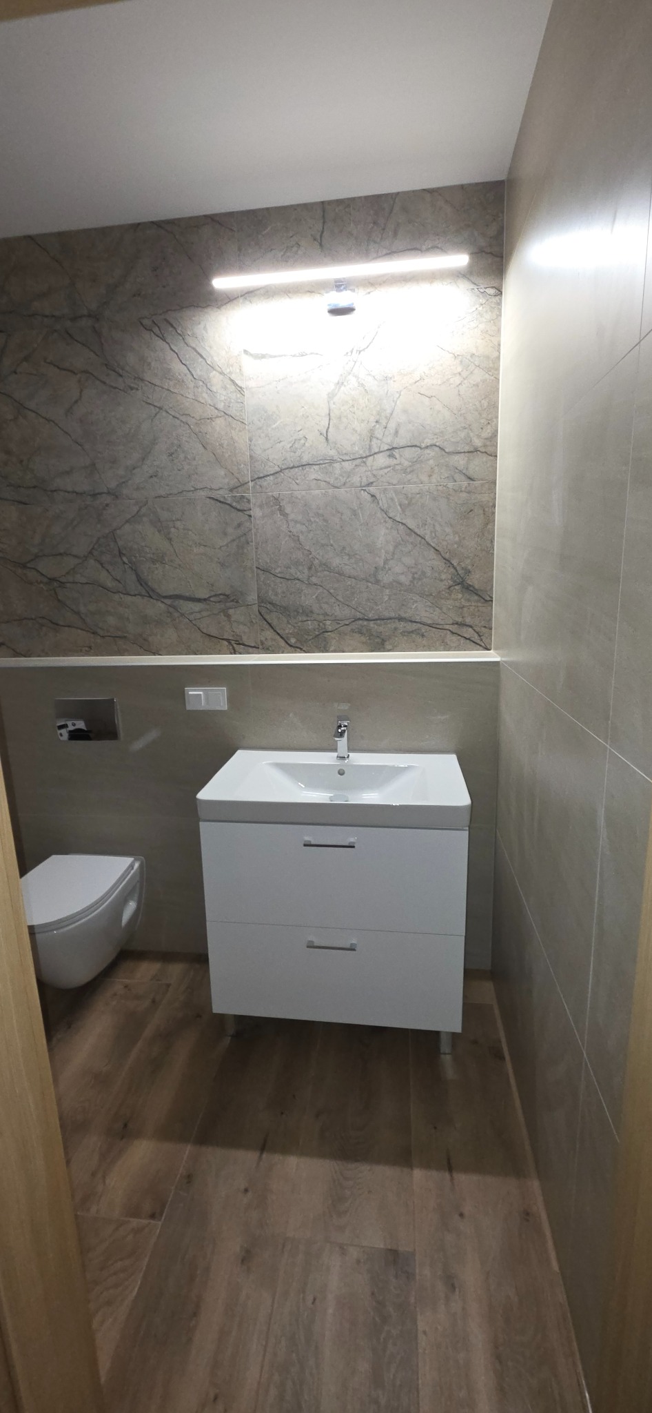 Nowoczesna łazienka po remoncie: biała szafka z umywalką, podwieszana toaleta, płytki imitujące marmur na ścianie i drewnopodobne na podłodze, oświetlenie LED.