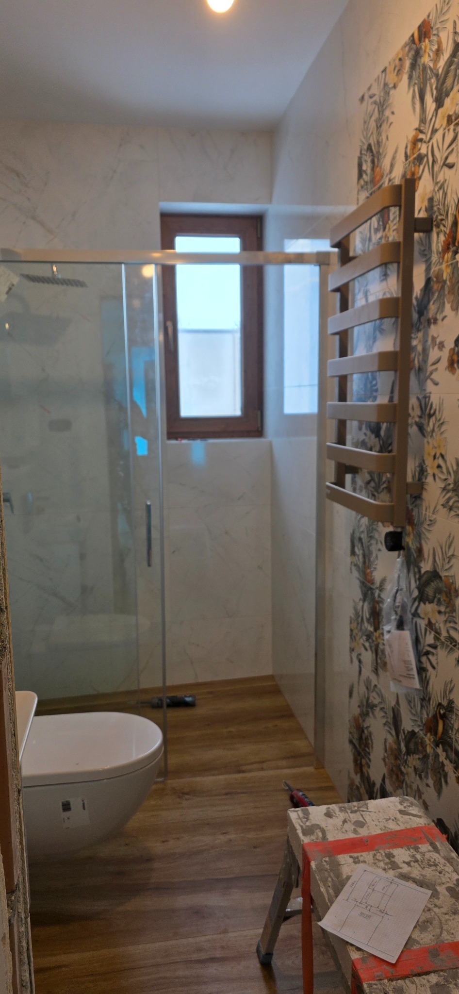 Wykończona łazienka z kabiną prysznicową, wiszącą toaletą, grzejnikiem drabinkowym na ścianie w kwiaty. Na podłodze panele imitujące drewno. Widoczny stołek roboczy.