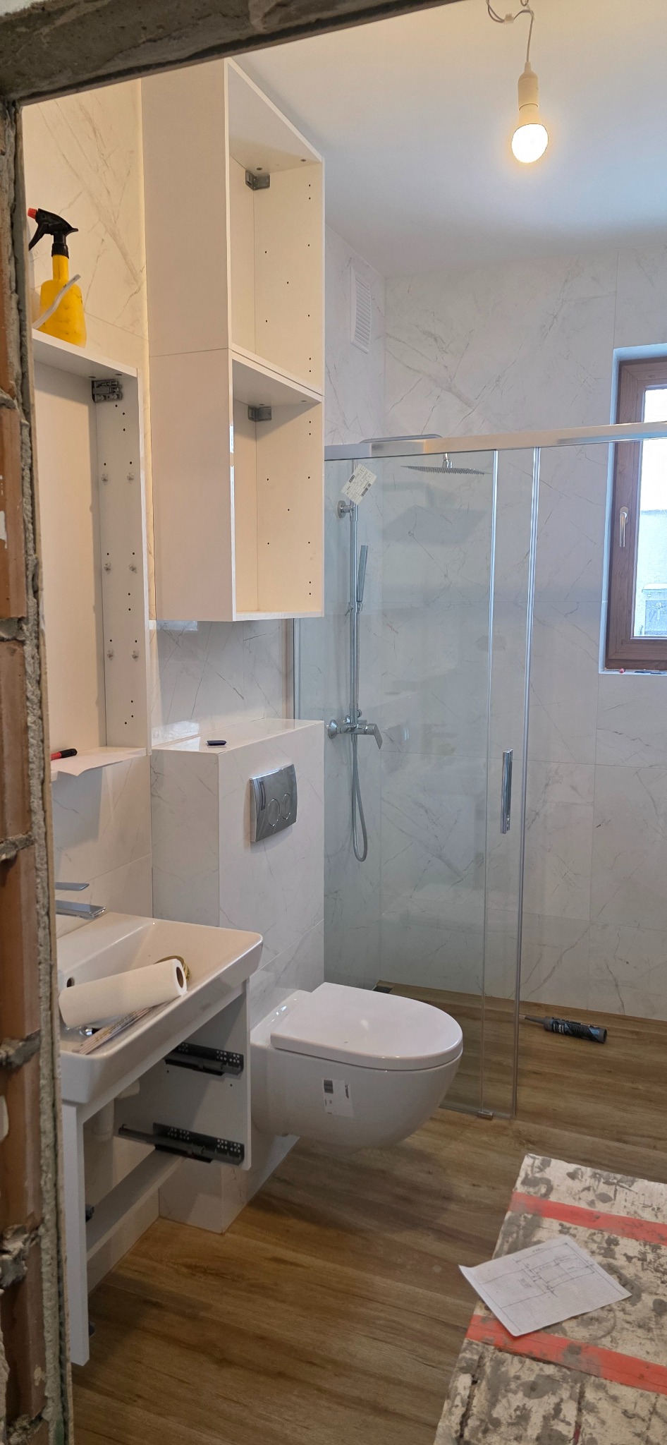 Nowoczesna łazienka w trakcie wykańczania: białe meble, kabina prysznicowa, drewnopodobne panele. Widoczne narzędzia i plany na podłodze. Oświetlenie górne.