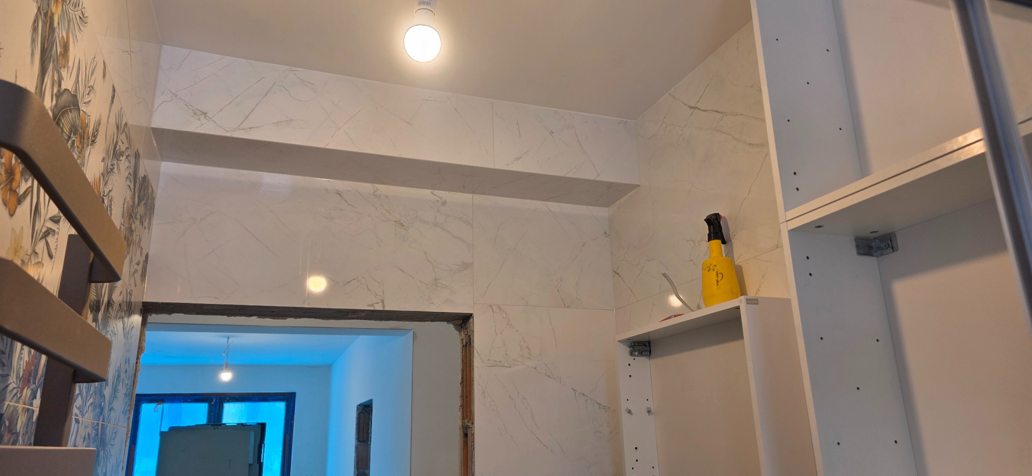 Wykończona łazienka z marmurowymi płytkami, widok na sąsiednie pomieszczenie. Żółty spryskiwacz na półce. Fragment szafki z widocznymi otworami montażowymi.