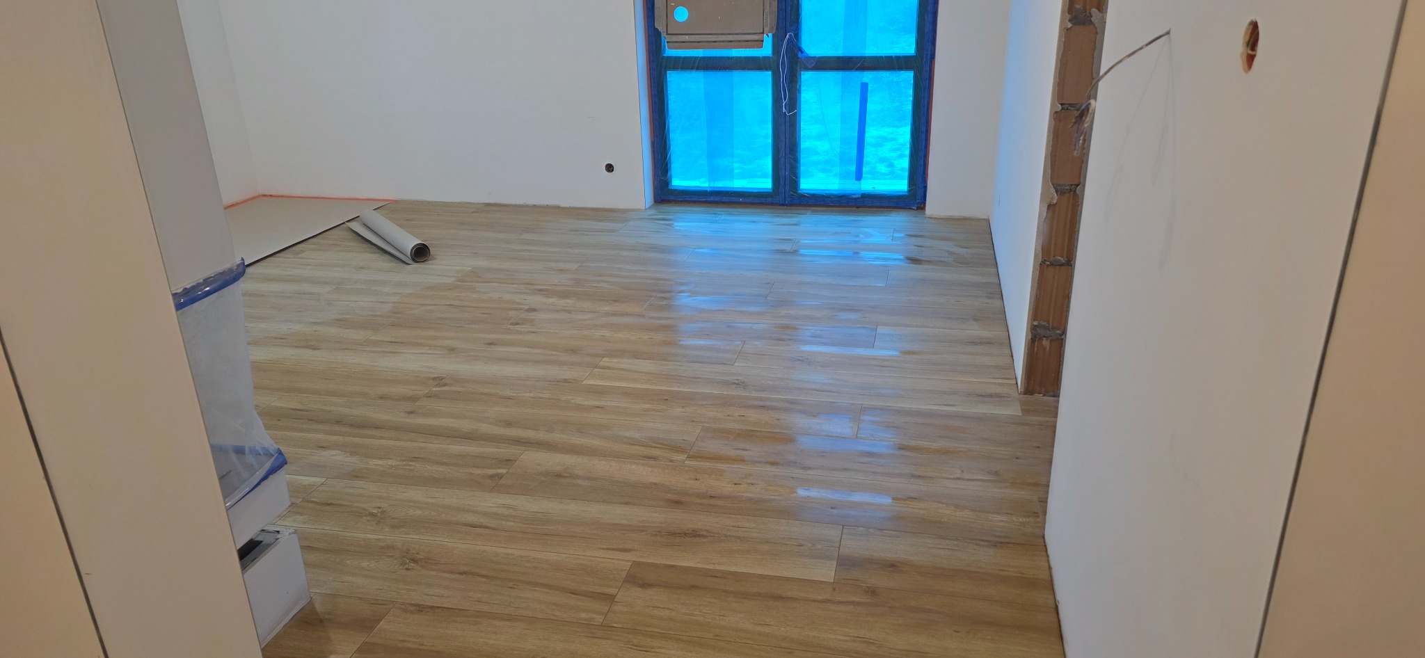 Ukończona podłoga z paneli imitujących drewno w jasnym wnętrzu. Okno zabezpieczone folią, widoczne przygotowania do dalszych prac wykończeniowych.