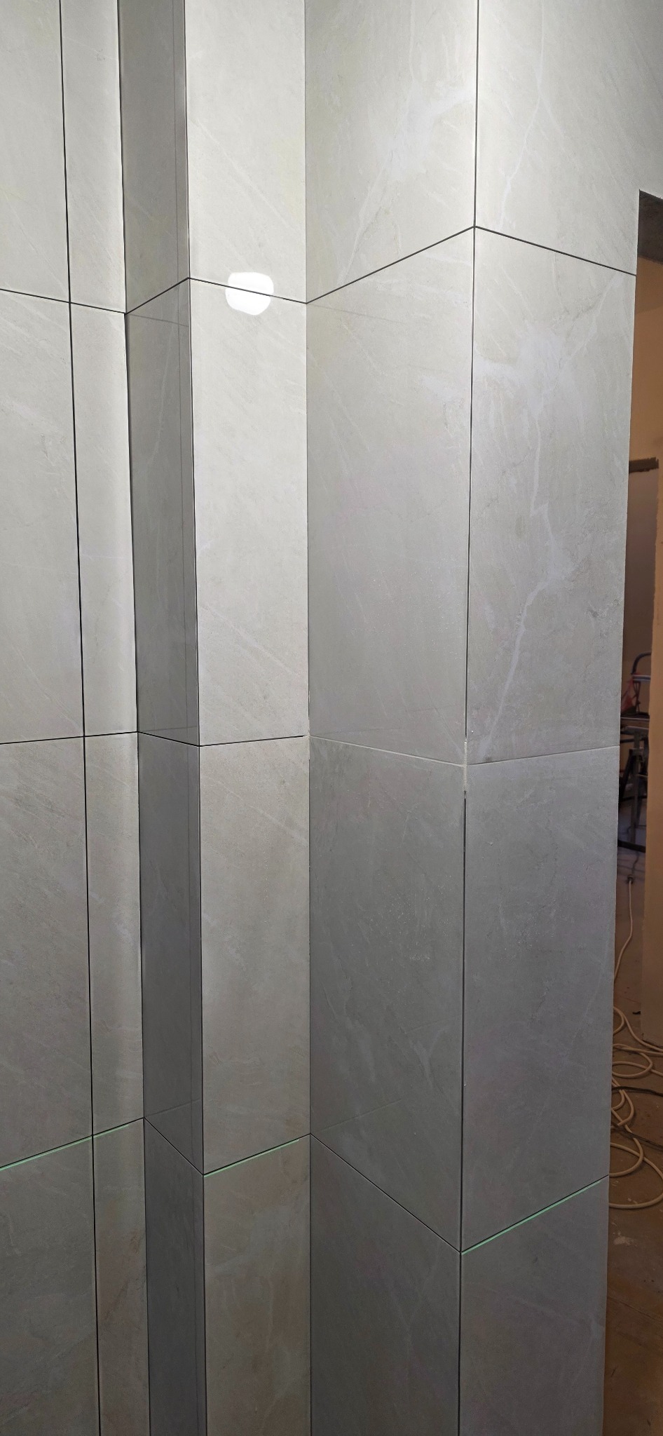 Ściana wyłożona jasnymi, marmurkowymi płytkami. Widoczne precyzyjne fugi i odbicie światła na powierzchni płytek. W tle fragment wnętrza w trakcie prac.