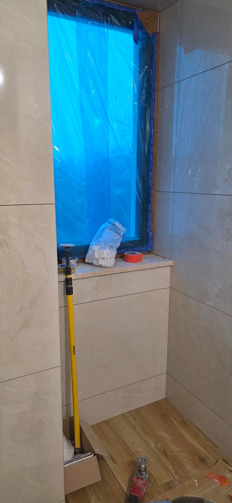 Układanie płytek w łazience w Chechło Pierwsze. Widoczne narzędzia glazurnicze i zabezpieczone okno folią. Jasne płytki na ścianach i podłodze.