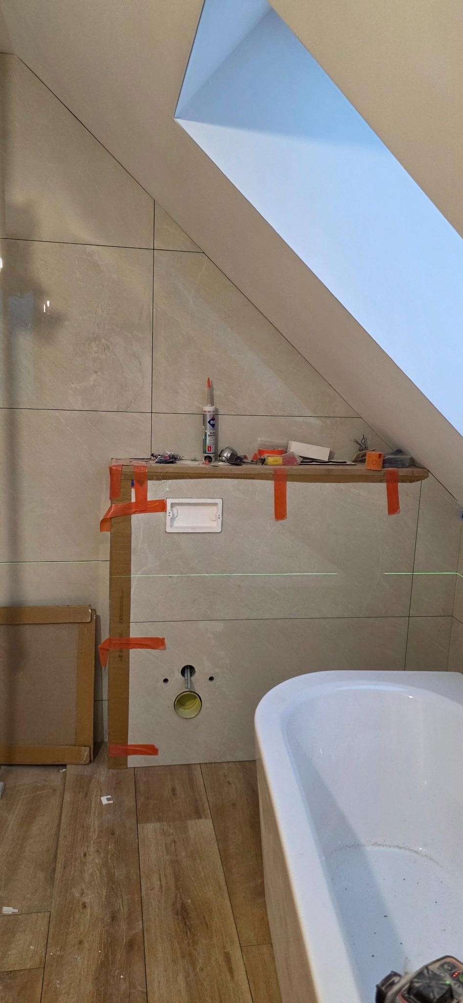Montaż płytek w łazience na poddaszu. Widoczne płytki imitujące kamień, wanna, laser krzyżowy i narzędzia. Prace w toku, zabezpieczenia z taśmy.
