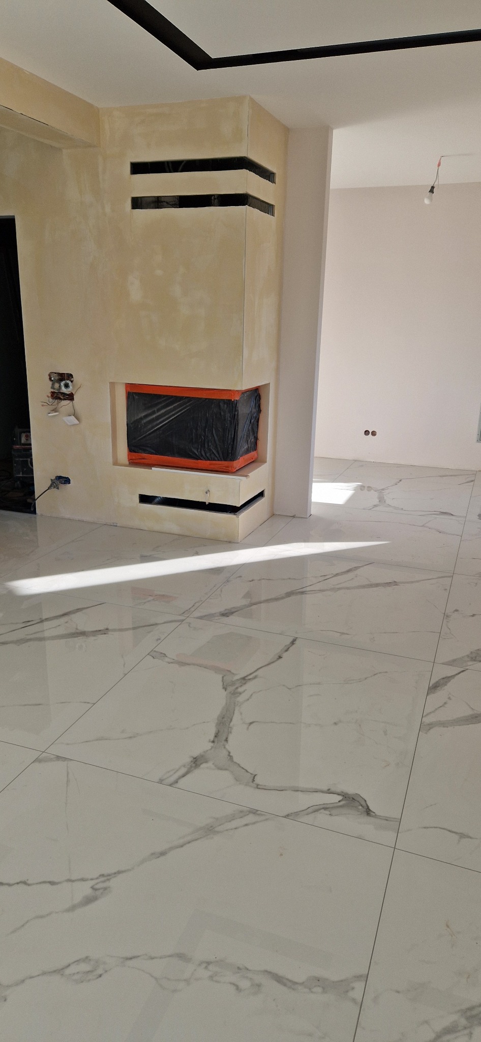 Wykończony kominek z gładzią w beżowym odcieniu, zabezpieczony folią. Na podłodze białe płytki imitujące marmur. Widoczne gniazdka elektryczne i fragment instalacji.