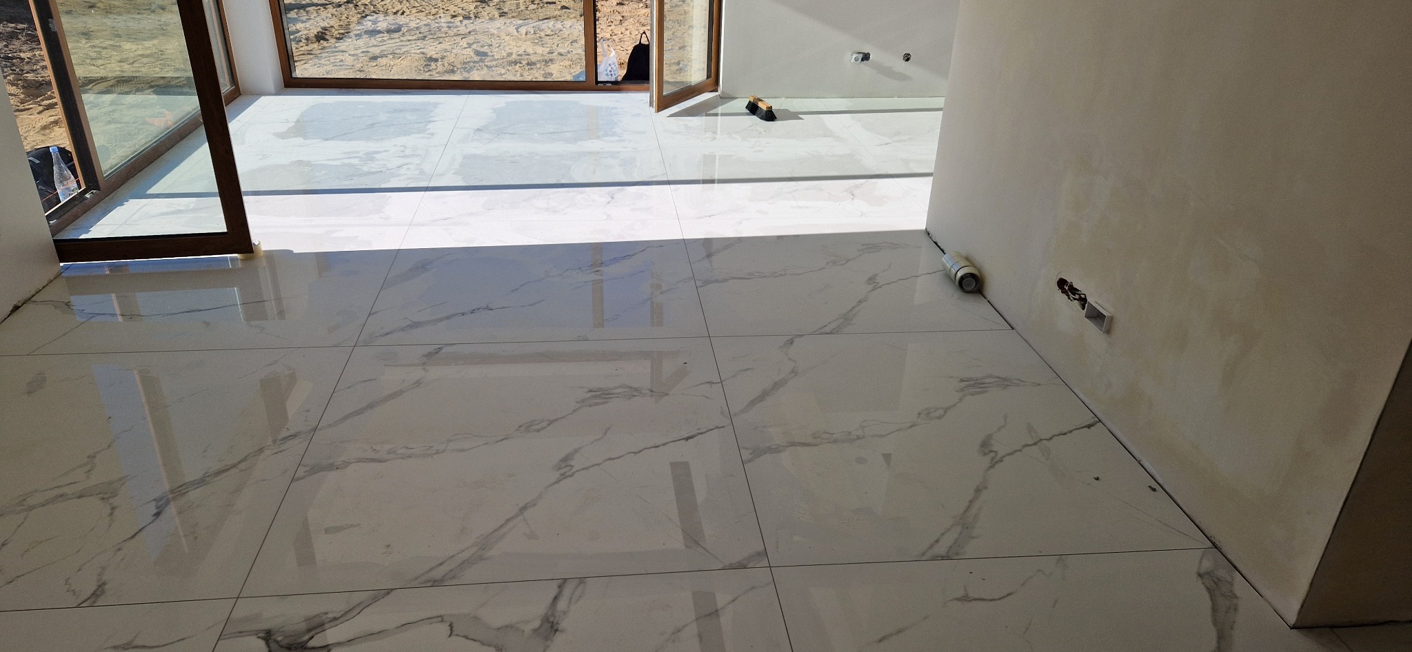 Błyszcząca, biała podłoga z płytek imitujących marmur w jasnym wnętrzu. Widoczne okno i fragment ściany z instalacją elektryczną.