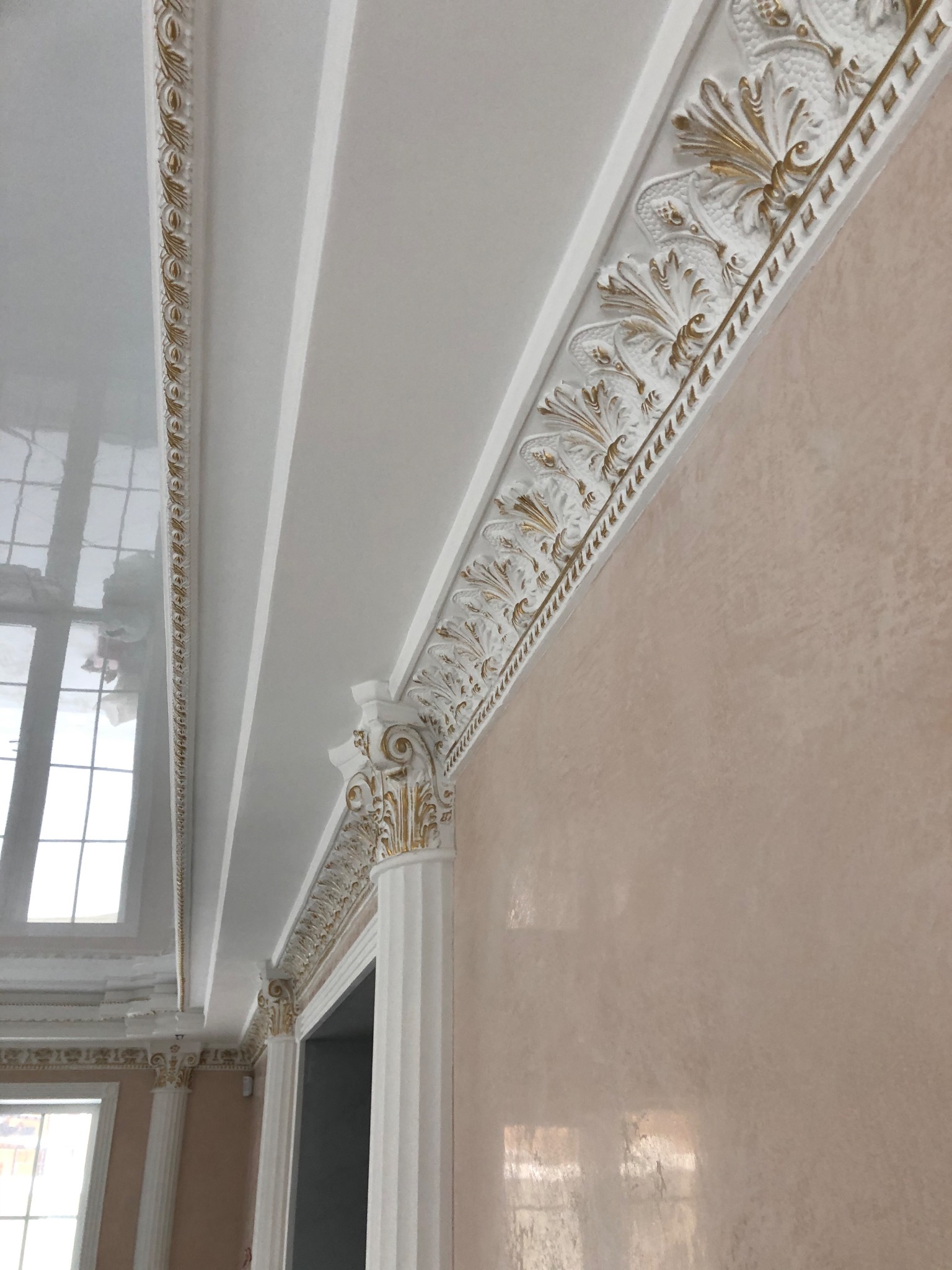 Wykończenie wnętrza z dekoracyjnym sztukaterią sufitową w kolorze białym ze złotymi akcentami, widoczne okno i ściana w odcieniach beżu, perspektywa od dołu.