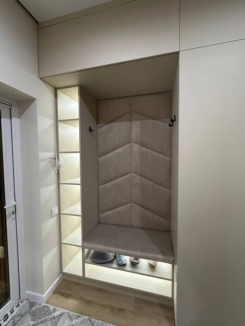 Nowoczesna zabudowa przedpokoju z tapicerowanym siedziskiem i podświetlanymi półkami, widok z bliska na estetyczne wykończenie i detale.