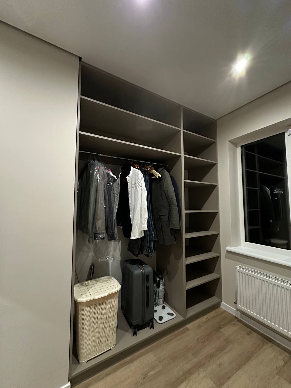 Szara, wnękowa szafa z przesuwnymi drzwiami i półkami, wypełniona ubraniami, walizką i koszem na pranie. Widok na okno i grzejnik w tle.