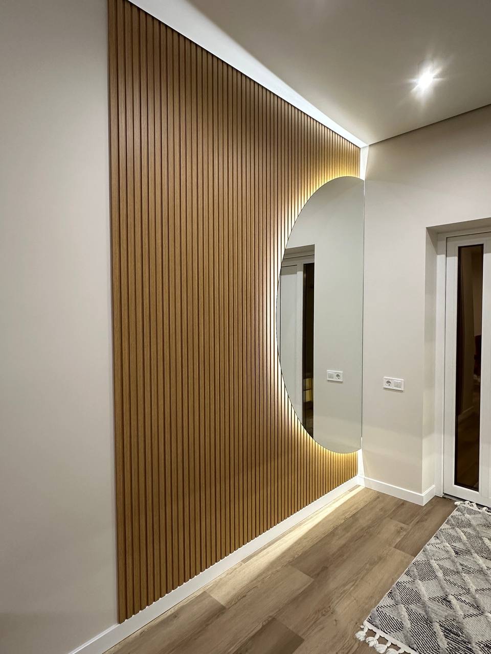 Nowoczesne wykończenie ściany lamelami drewnianymi z okrągłym lustrem podświetlonym LED, w jasnym wnętrzu. Widok z boku, podłoga drewnopodobna.