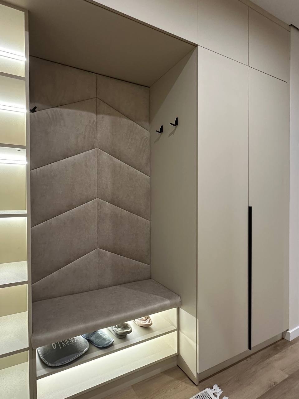 Stylowa zabudowa przedpokoju z tapicerowanym siedziskiem i panelami ściennymi w kształcie strzałek, oświetlona listwami LED, z szafą w neutralnej kolorystyce.