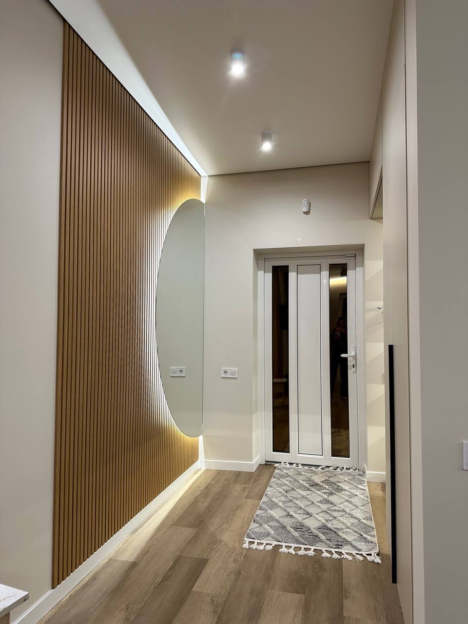 Nowoczesny przedpokój z drewnianą ścianą, okrągłym lustrem z podświetleniem LED i jasną podłogą. Minimalistyczny design, białe ściany, dywanik w geometryczne wzory przy drzwiach.