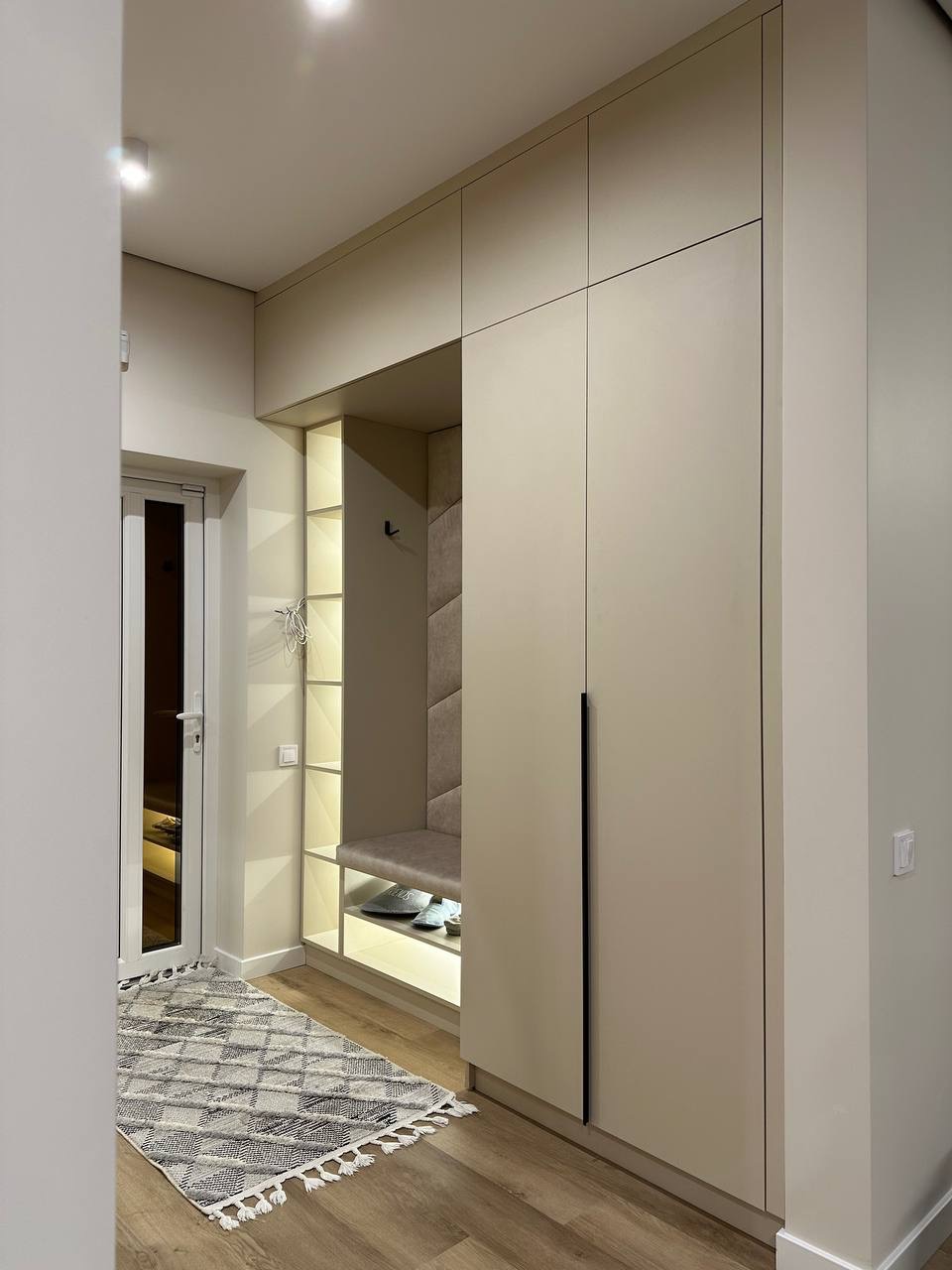 Beżowa zabudowa przedpokoju z siedziskiem i oświetleniem LED. Minimalistyczny design, drewnopodobna podłoga i dywanik w romby. Nowoczesne wykończenie wnętrza.
