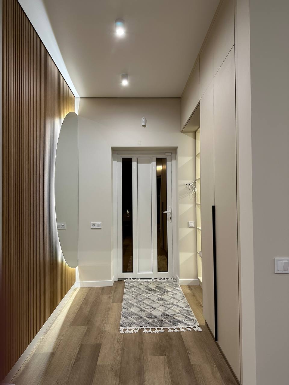 Nowoczesny przedpokój z drewnianą boazerią, okrągłym lustrem z podświetleniem i białymi szafami. Jasne drewno na podłodze i geometryczny dywan.