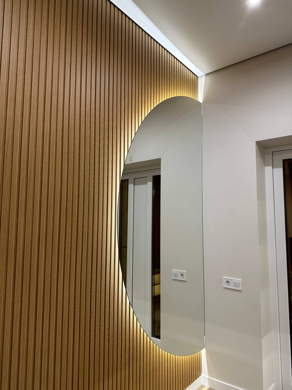 Nowoczesne wykończenie wnętrza: drewniane panele ścienne z podświetleniem LED i owalne lustro. Widoczne gniazdka elektryczne i fragment drzwi.