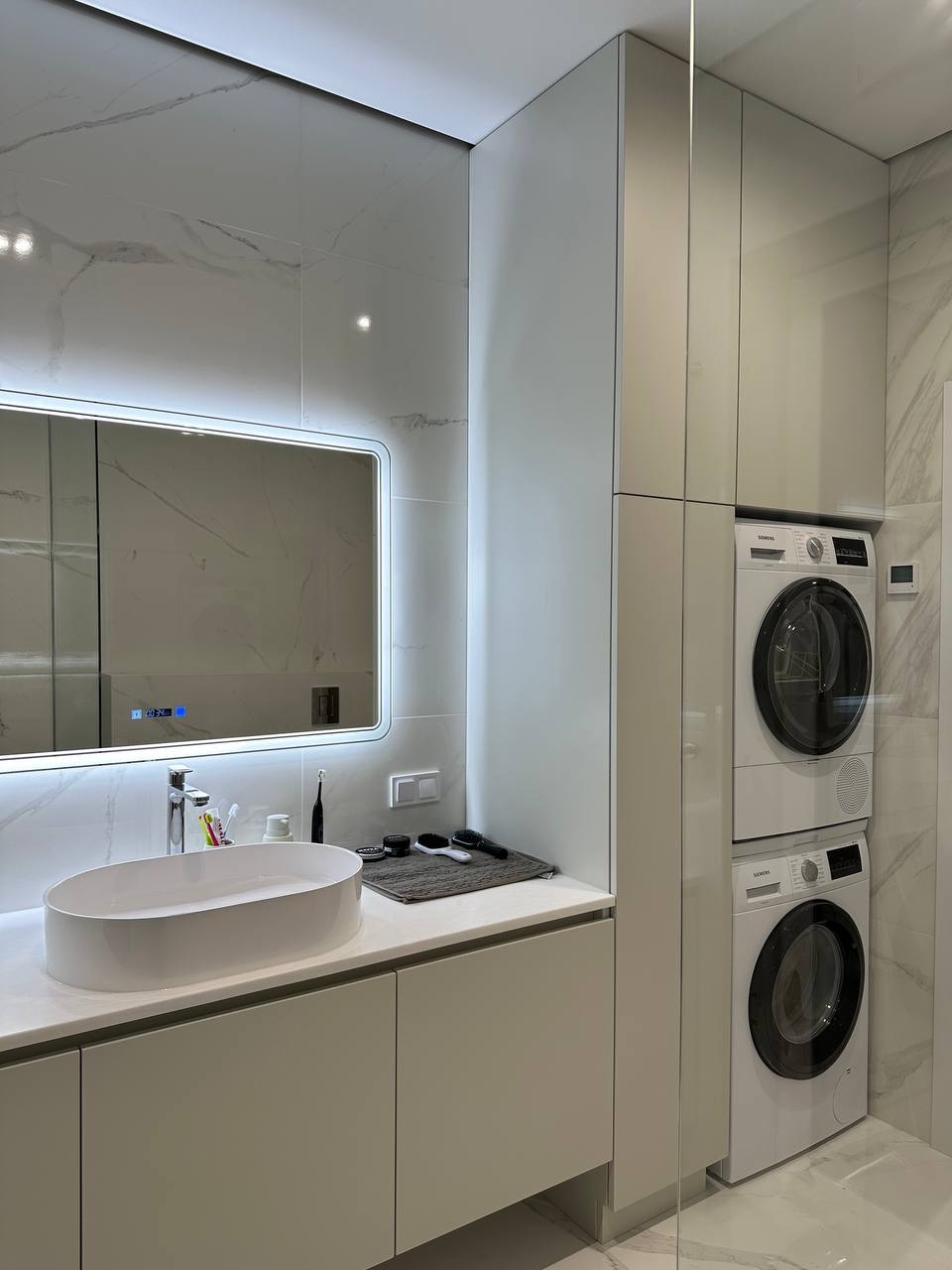 Nowoczesna łazienka z umywalką nablatową, podświetlanym lustrem i pralko-suszarką w zabudowie. Marmurowe płytki na ścianach dodają elegancji.