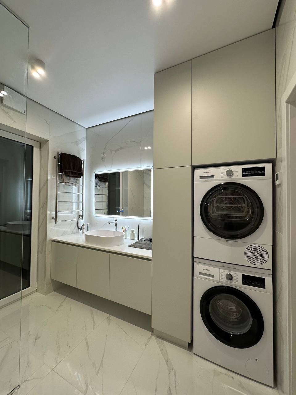 Nowoczesna łazienka z marmurowymi płytkami, okrągłą umywalką i zabudowaną pralko-suszarką Siemens. Minimalistyczny design z podświetlanym lustrem.
