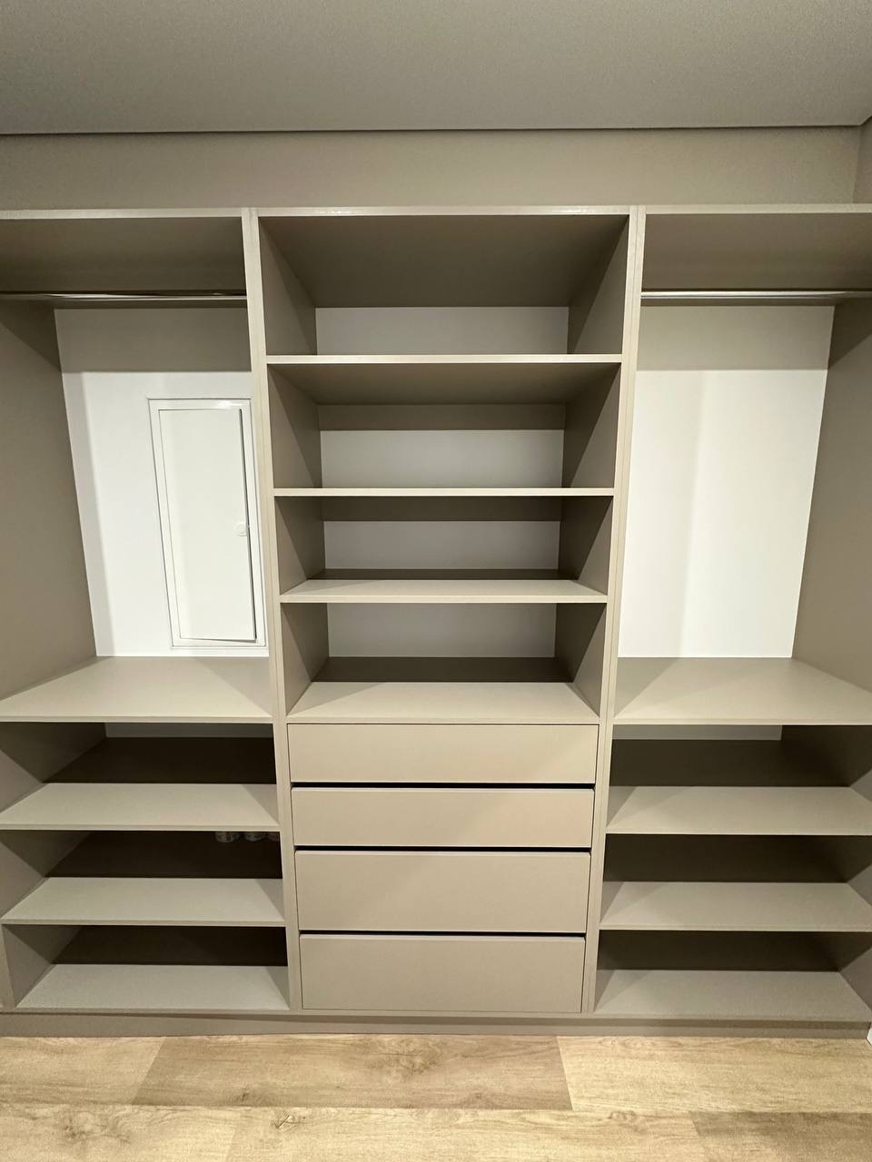 Beżowa, zabudowana szafa z półkami i szufladami, widoczna z przodu. Minimalistyczny design, jasne drewno na podłodze. Ukończona realizacja.