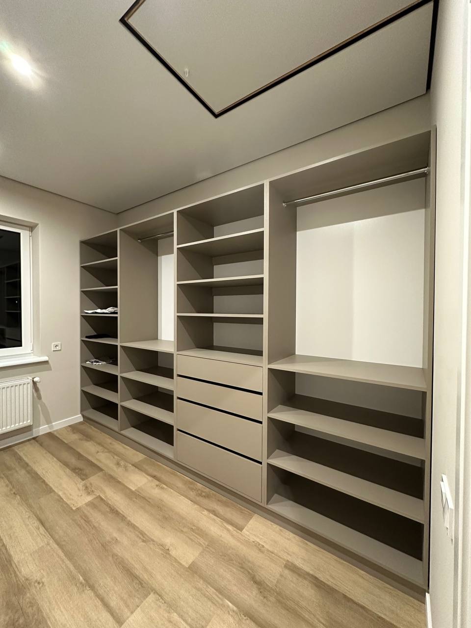 Jasna garderoba z szufladami i półkami, drewniana podłoga. Minimalistyczny design, widok na okno z grzejnikiem. Neutralna kolorystyka, nowoczesne wykończenie.