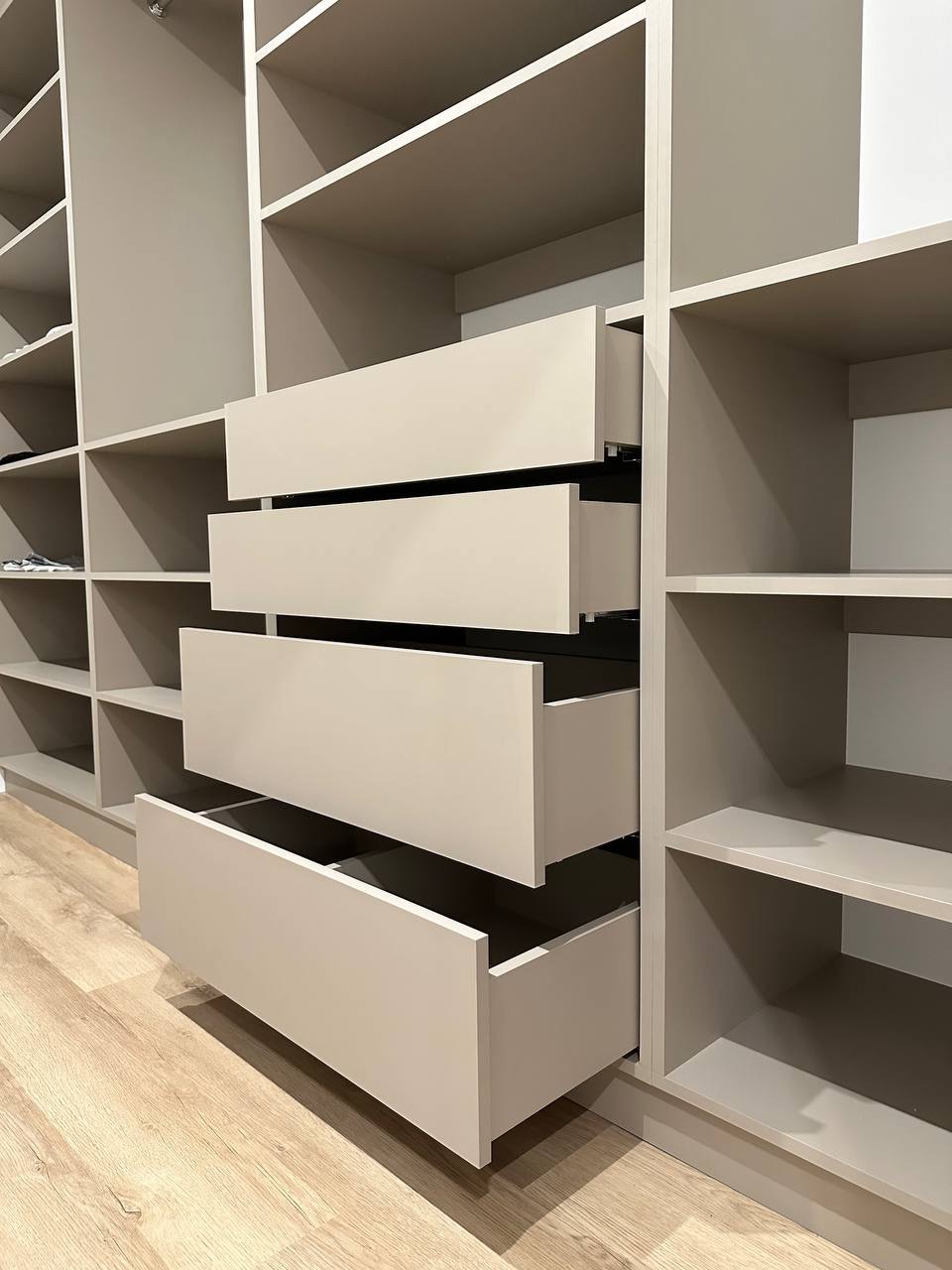 Garderoba z szufladami w odcieniach beżu, częściowo wysunięte. Widoczna drewniana podłoga i półki. Całość utrzymana w minimalistycznym stylu.