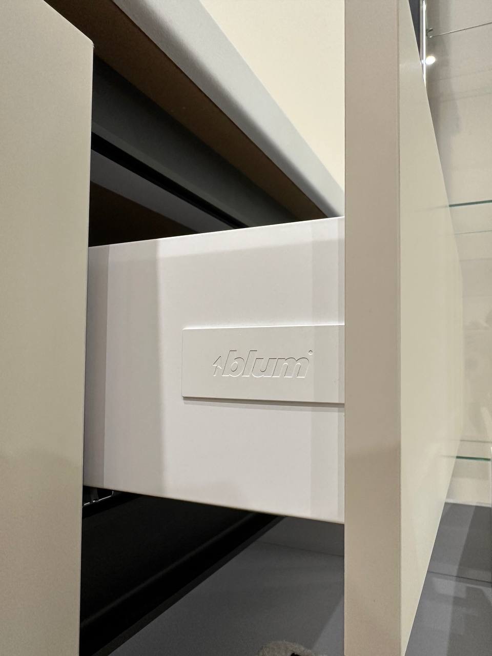 Biała szuflada z logo Blum, częściowo wysunięta, z widocznymi elementami prowadnic i wnętrza szafki. Minimalistyczny design, neutralne tło, oświetlenie dzienne.